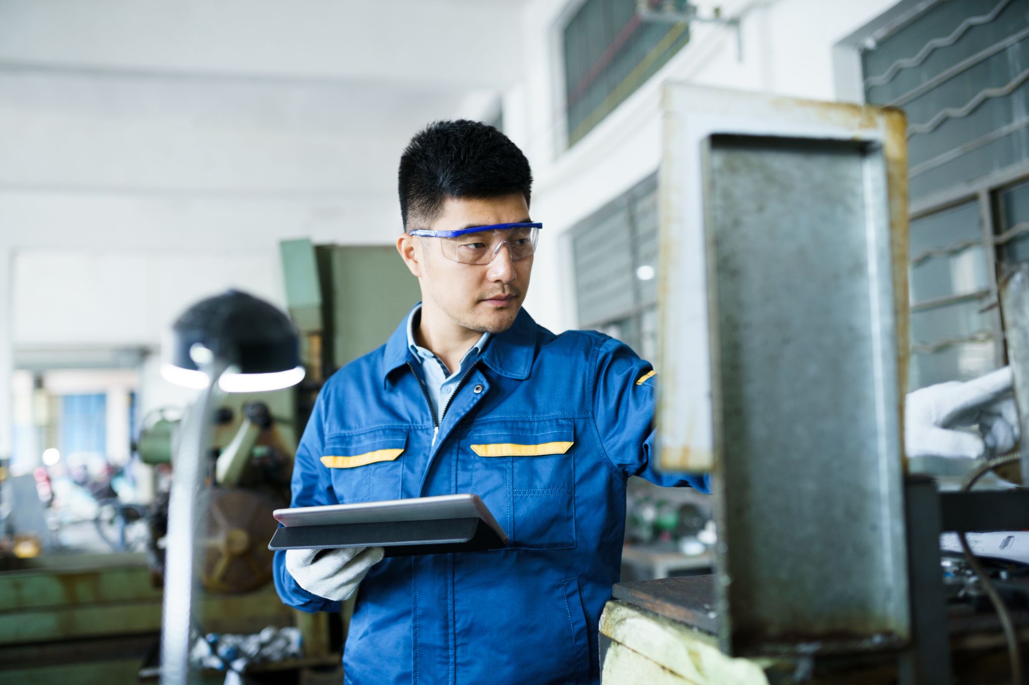 Image of a person wearing a uniform and safety goggles, working on a machine, and holding a tablet.